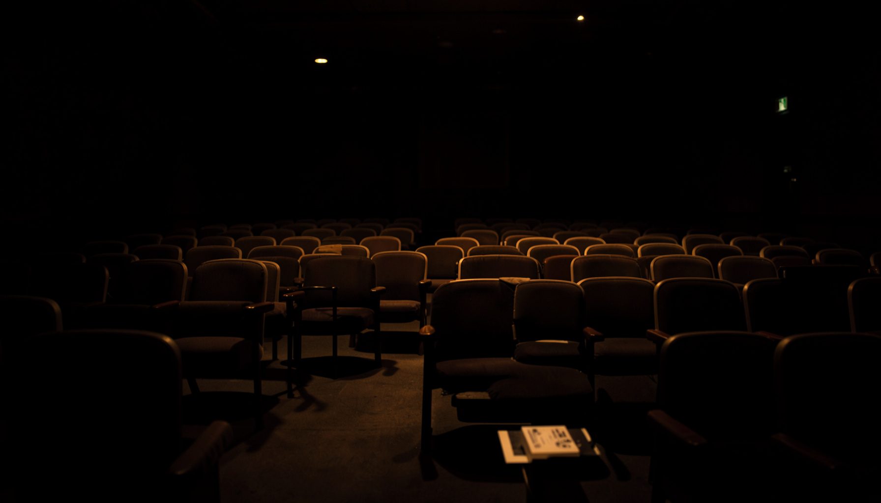 A dark, atmospheric photograph of a formal conference hall with rows of upholstered chairs, low warm lighting, and deep shadows across the seating area, conveying a quiet institutional setting.